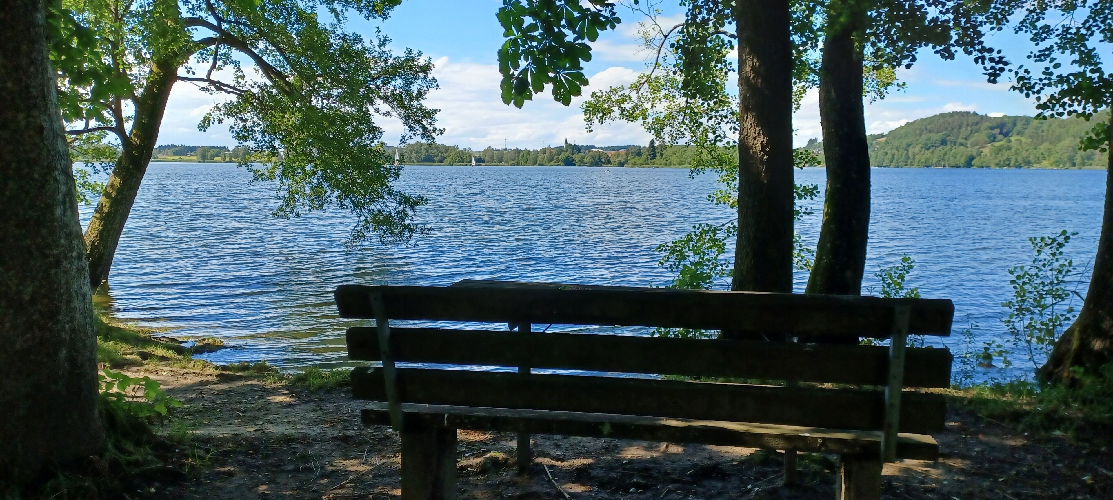 Puinen penkki järven rannalla, taustalla näkyy puita ja kukkuloita.