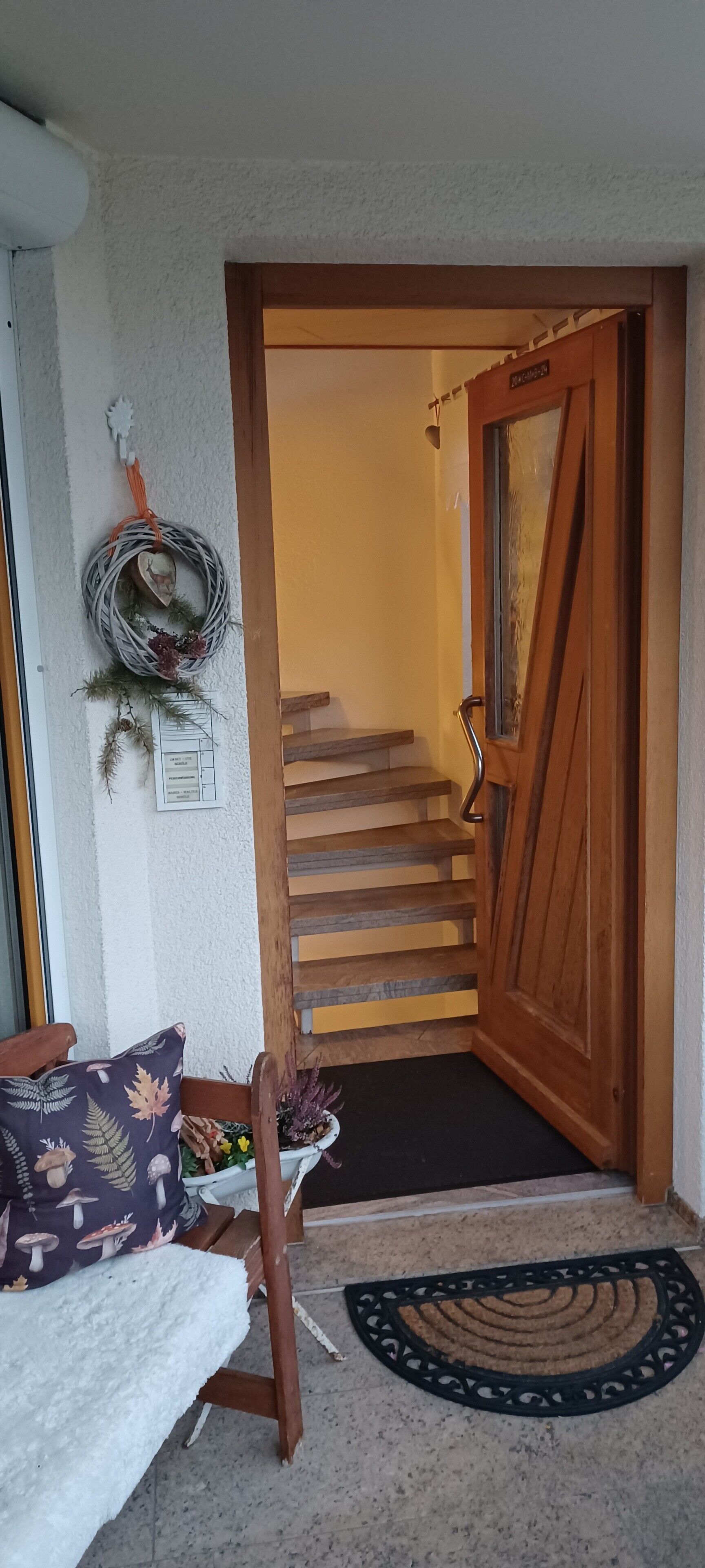 Eine Treppe führt zu einem Raum, eine Holztür, eine weiße Wand mit einem Kranz, ein Stuhl mit einem gemusterten Kissen und eine Fußmatte.