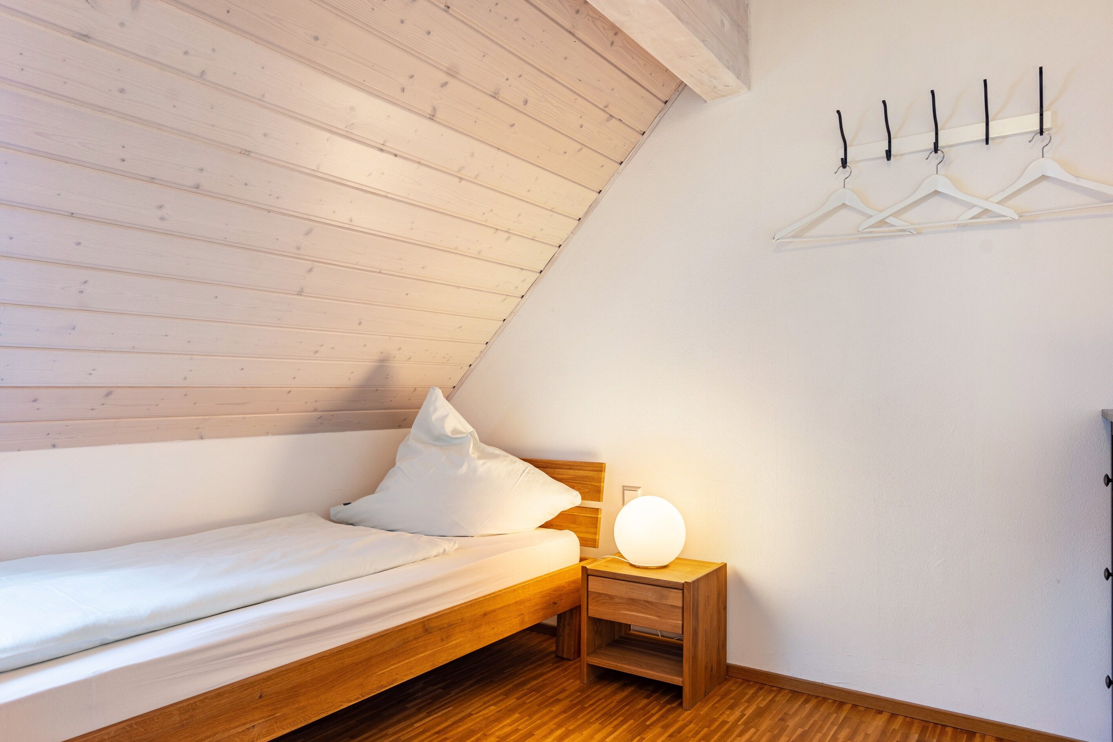 Un lit simple avec une tête de lit en bois, une table de chevet avec une lampe et un porte-manteau fixé au mur.