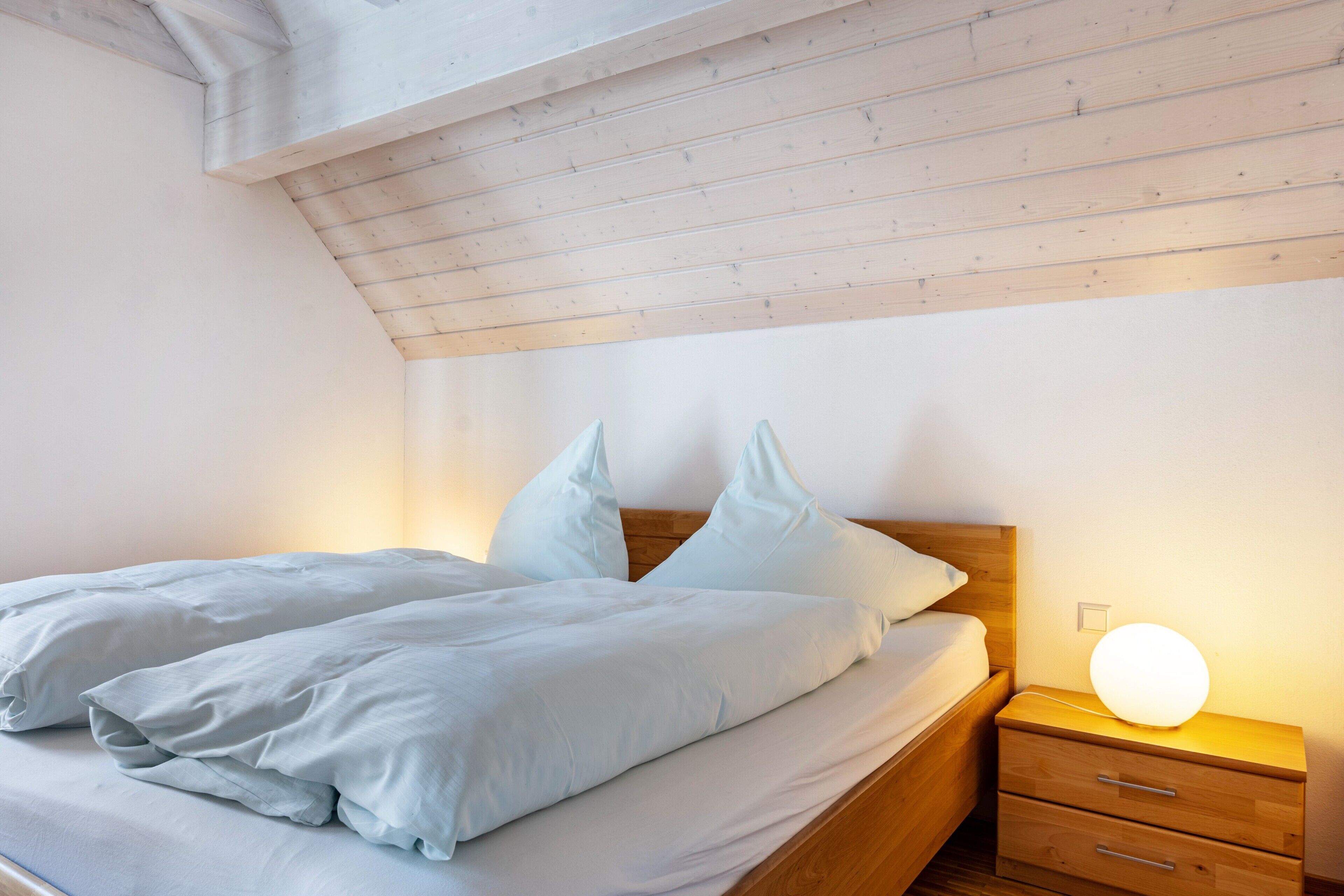A bedroom with a wooden headboard, a bedside table with a lamp, and a bed with white bedding.