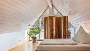 Un dormitorio con techo inclinado, una cama con ropa de cama blanca, una planta en maceta y un separador de madera.