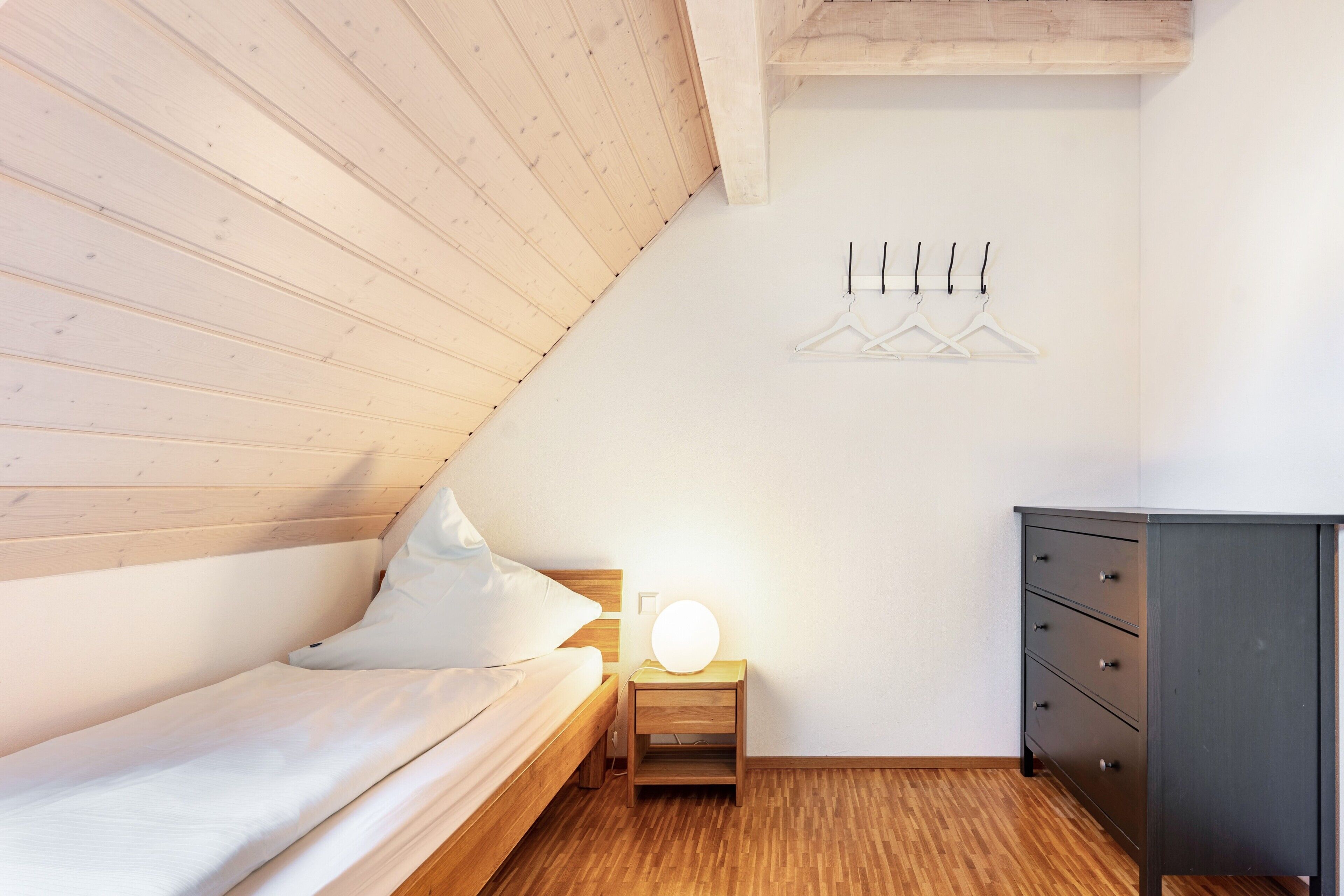 Ett sovrum med ett träpanelat tak, en säng med vita sängkläder, ett sängbord med en lampa, en byrå och en väggmonterad klädhängare.