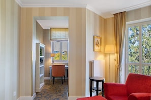 Quarto de hotel com uma poltrona vermelha, uma mesa pequena, uma luminária, uma janela com vista para árvores e uma porta que dá para uma sala de jantar.