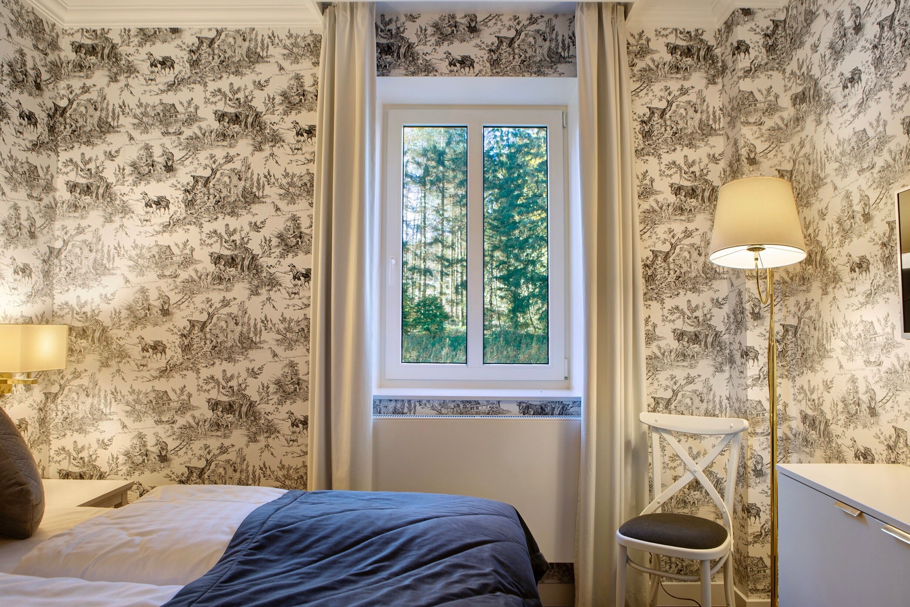 A bedroom with a bed, a chair, a lamp, and a window with a view of trees.