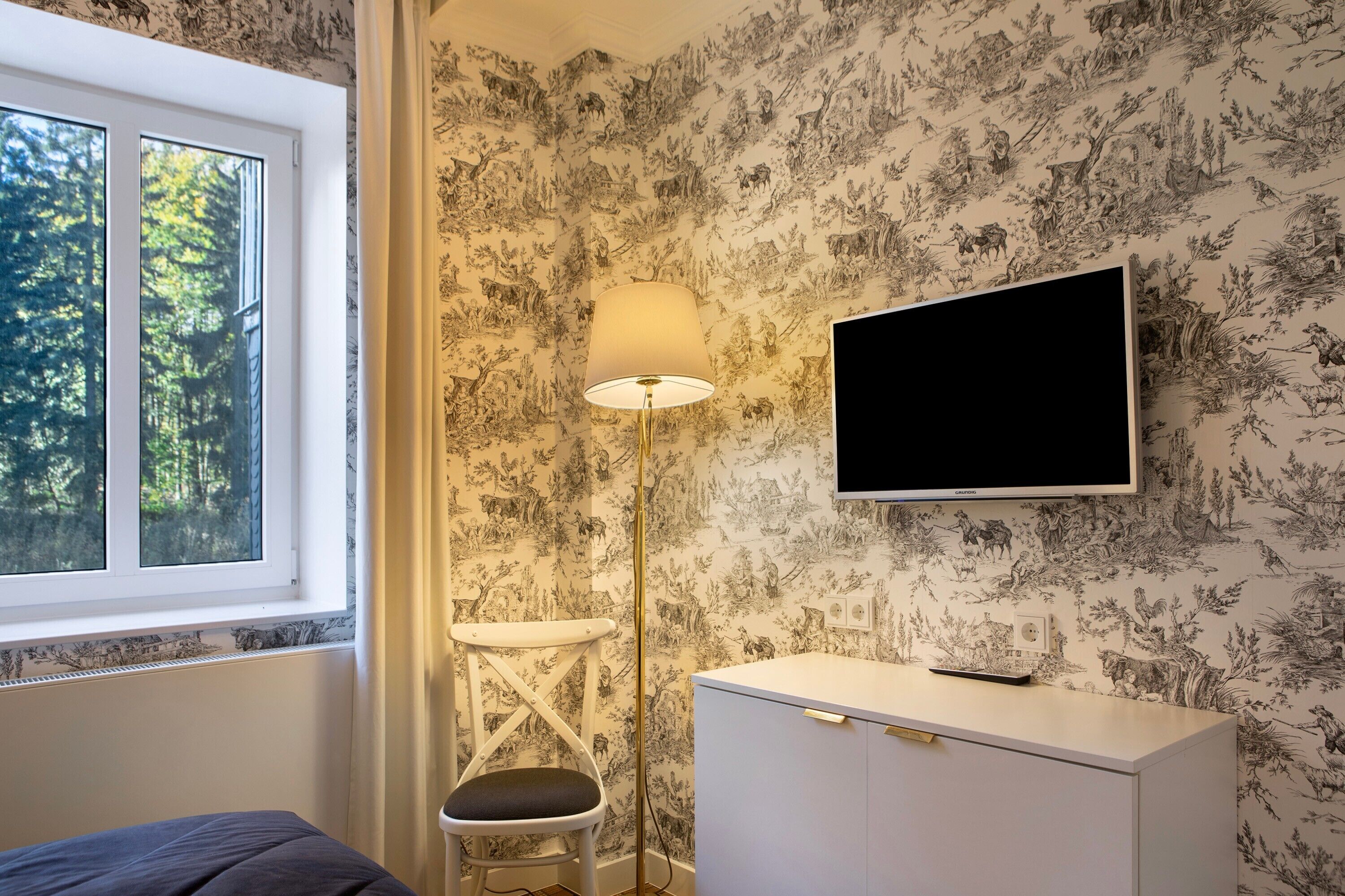 Quarto com cama, escrivaninha, cadeira, luminária, televisão e janela com vista para árvores.