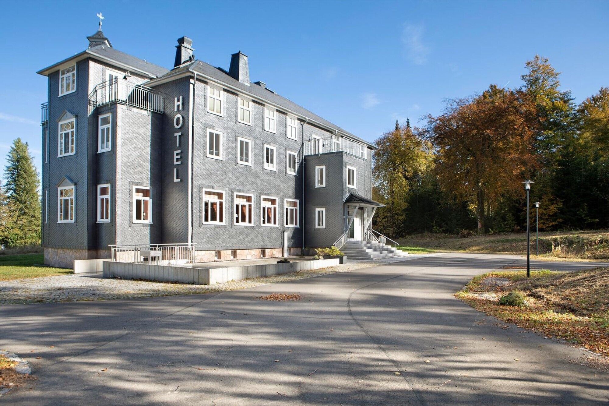 A large, multi-story hotel with a prominent 'HOTEL' sign on the facade, a central entrance with a staircase, and a driveway leading to the entrance.