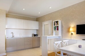 A modern kitchen with white cabinets, a built-in bookshelf, a flat-screen TV, and a dining area with chairs.