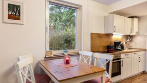 A kitchen with a wooden table, white chairs, a window with a view of trees, and a small stove. - Apartment “Island of Relaxation Bungalow” with Private Terrace, Garden & Wi-Fi (Milmersdorf)