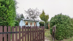 A small house with a light blue exterior, surrounded by a wooden fence and dense greenery. - Apartment “Island of Relaxation Bungalow” with Private Terrace, Garden & Wi-Fi (Milmersdorf)