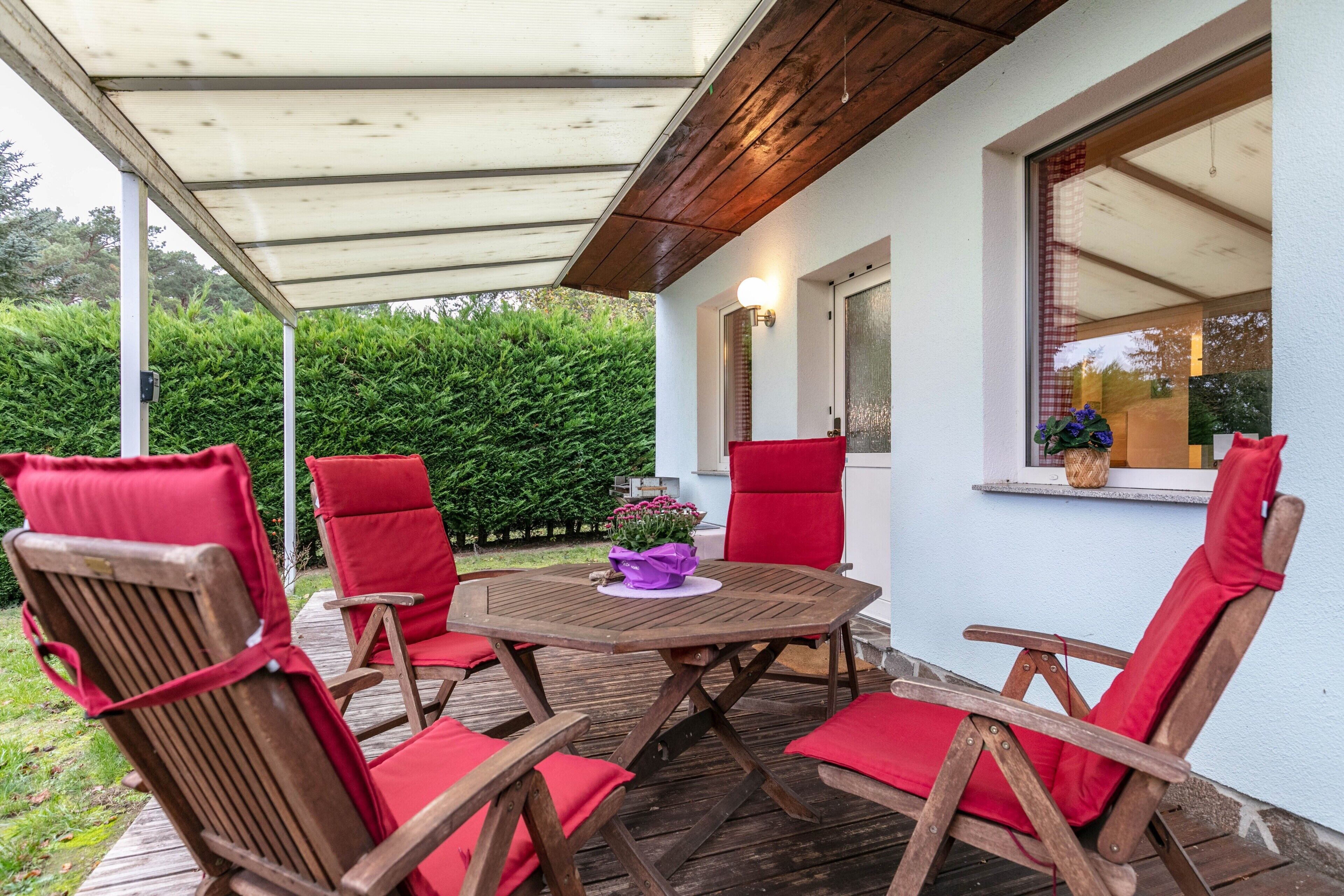 Eine überdachte Terrasse mit Holzsesseln und einem Tisch, ein Fenster mit Blick auf Grünflächen und eine Wandleuchte.