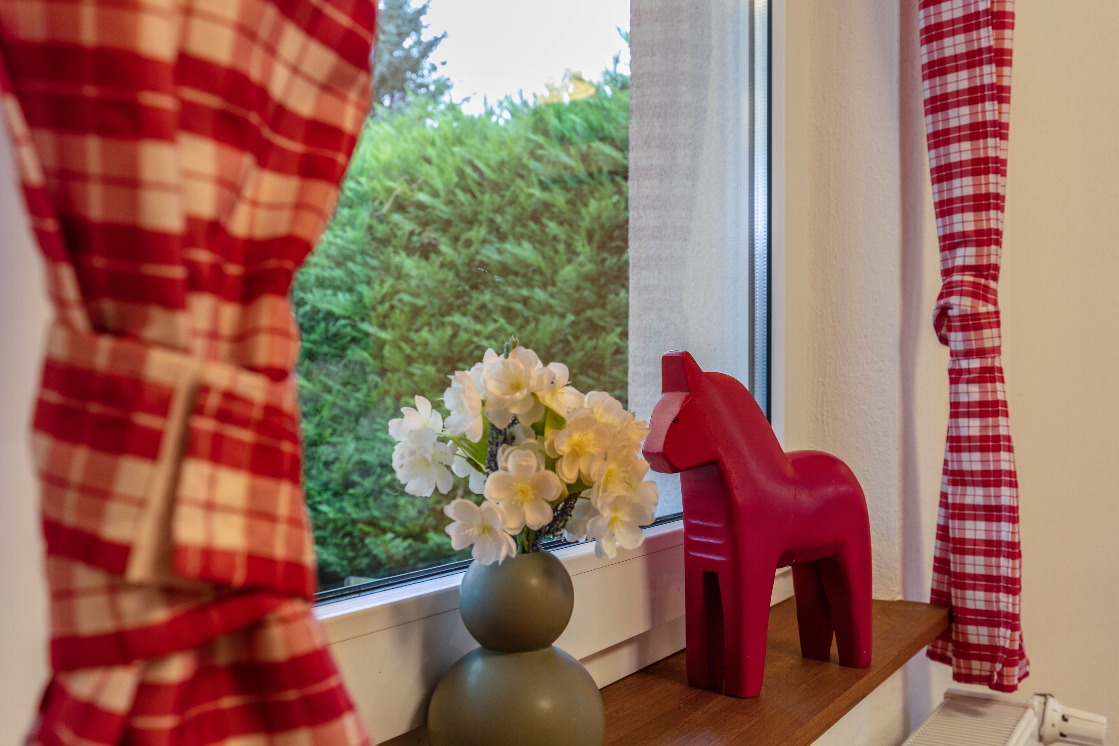 Une fenêtre avec un vase de fleurs, un objet rouge en forme de cheval et un rideau à carreaux.