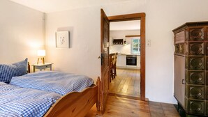 Un dormitorio con cama de madera, una mesita de noche con lámpara y vista a una cocina por una puerta abierta.
