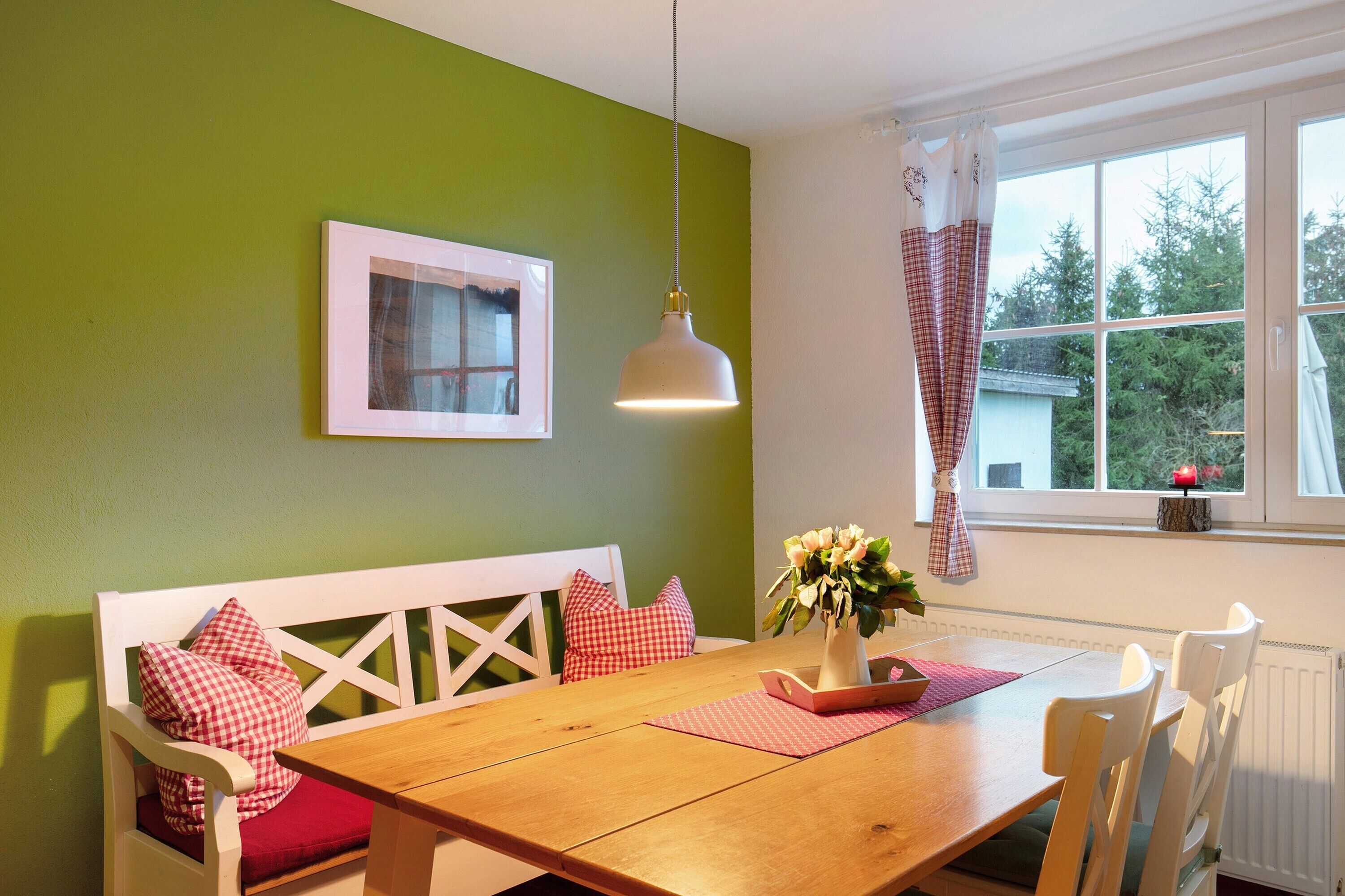 Een eetruimte met een houten tafel, witte stoelen, een bank met rode kussens, een hangende lamp, een ingelijst kunstwerk aan de muur en een raam met uitzicht op bomen.