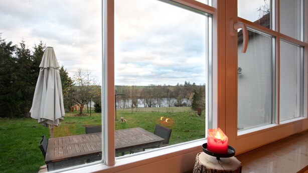 A room with a large window, a wooden table, and a candle on a tree stump. - Apartment 'Haus Am See - Zur Roten Tür' with Lake View, Private Terrace and Wi-Fi (Templin)