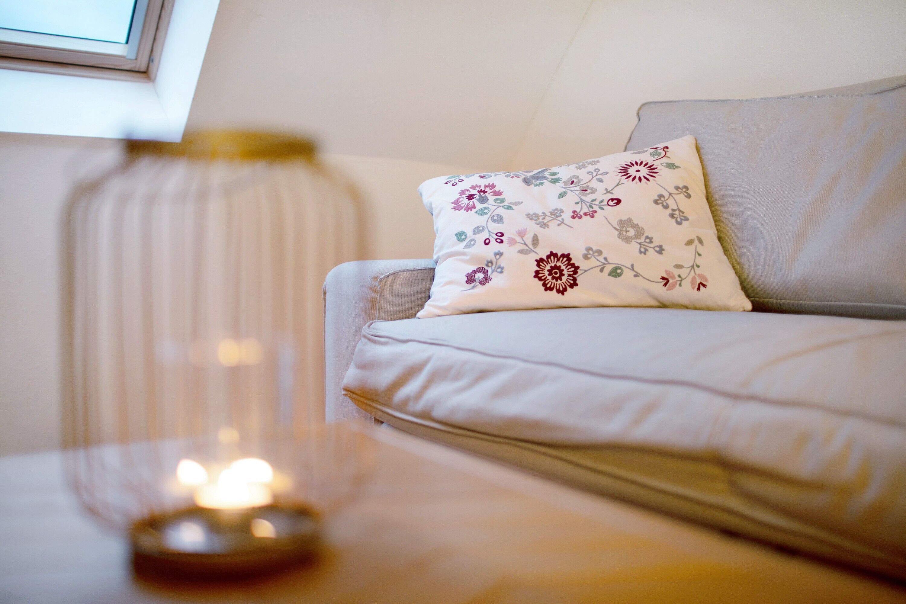 Une pièce avec un canapé blanc, un coussin à motifs floraux, une bougie sur une table et une fenêtre avec des stores.