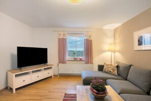 A living room with a flat-screen TV, a sofa, a coffee table with a flower vase, a lamp, and a window with curtains.