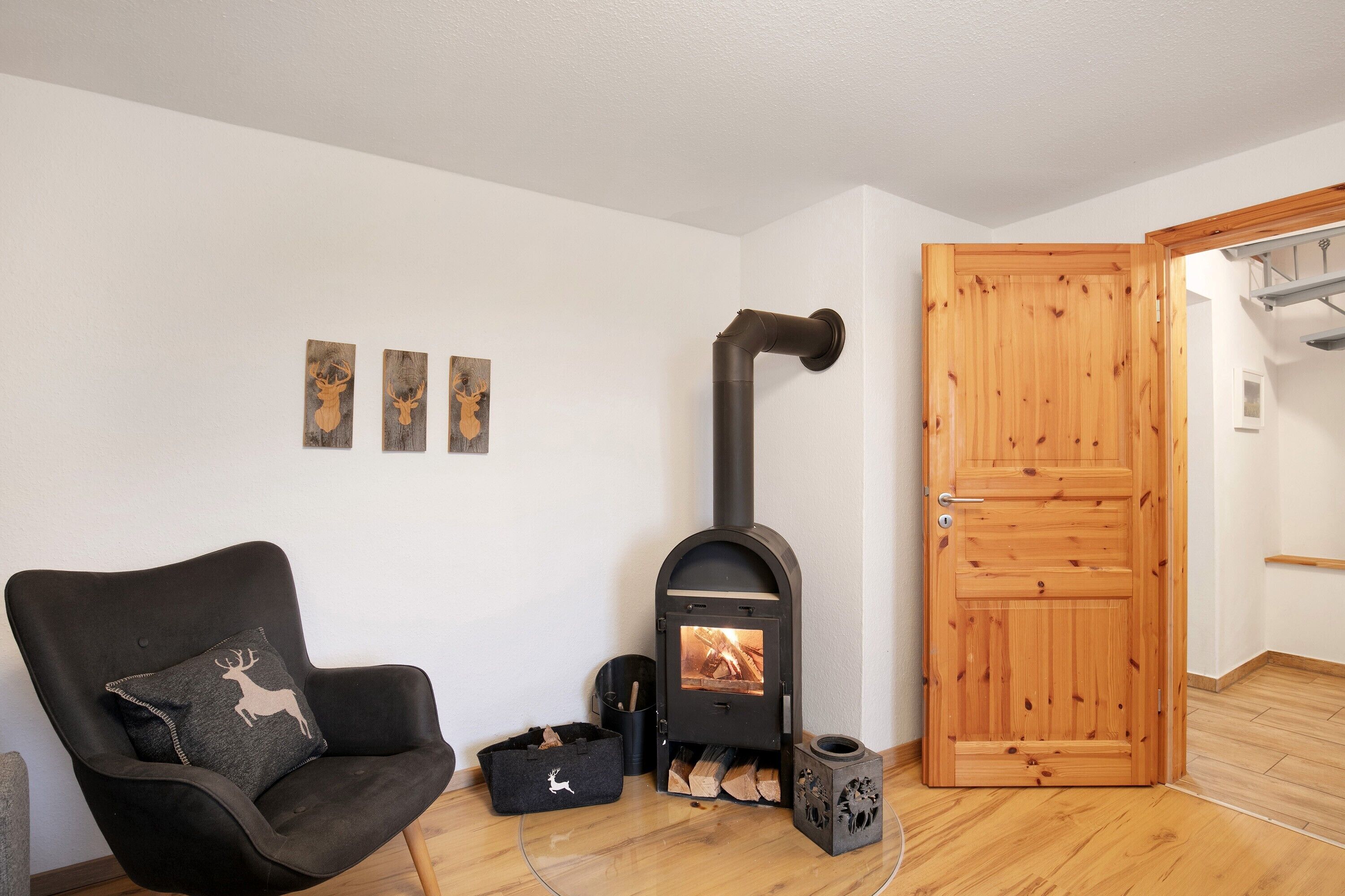 A room with a black upholstered armchair, a wood-burning stove, a wooden door, and three wall-mounted deer-themed decorations.