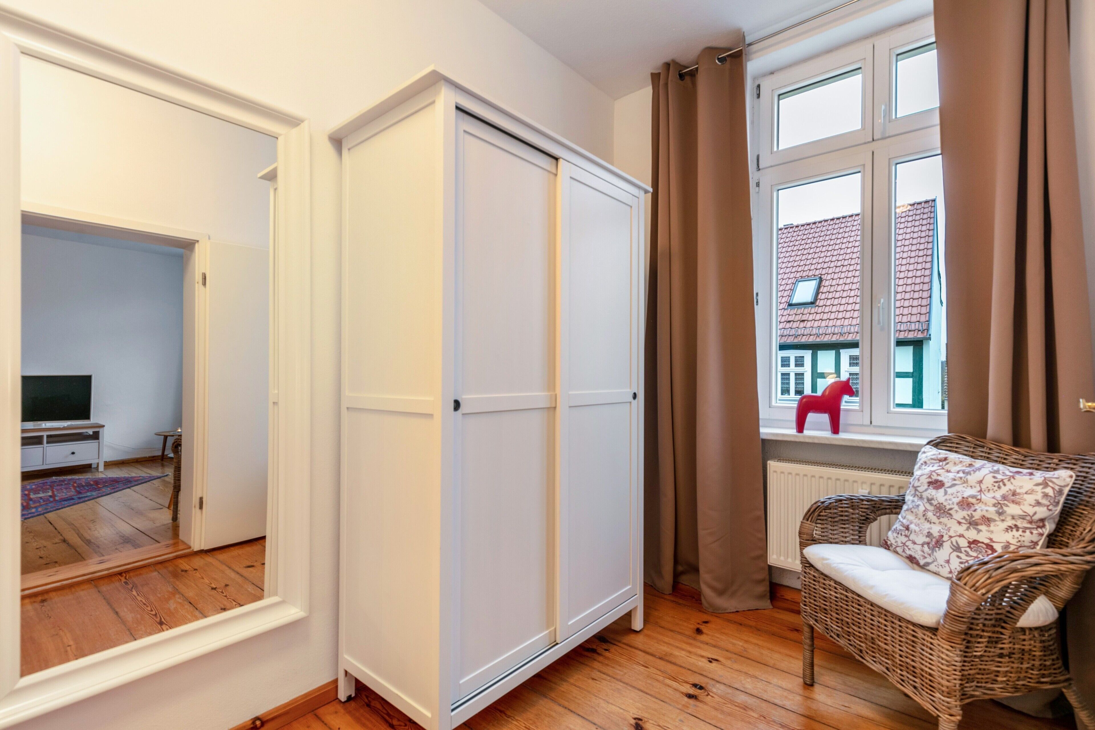 A room with a wardrobe, a wicker chair, a window with curtains, and a radiator.
