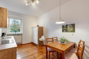 Uma cozinha compacta com armários de madeira, geladeira branca, mesa de jantar com cadeiras, luminária pendente e janela com vista para árvores.