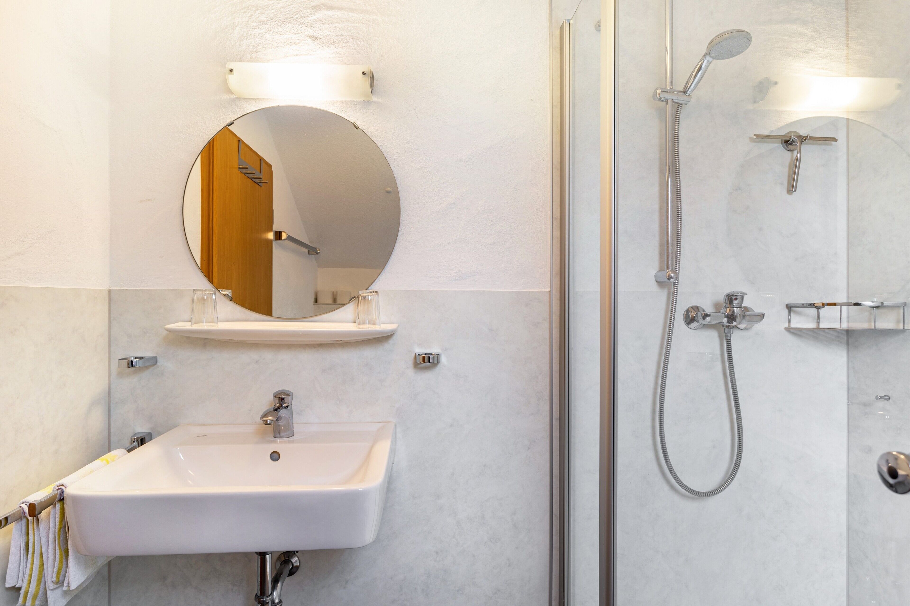 A bathroom with a white sink, a round mirror, a shower with a handheld showerhead, and a glass-enclosed shower stall.