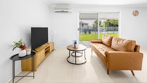 Ein modernes Wohnzimmer mit Flachbildfernseher, Holzschrank, rundem Couchtisch, beiger Couch und einer großen Schiebeglasdoor zum Balkon.