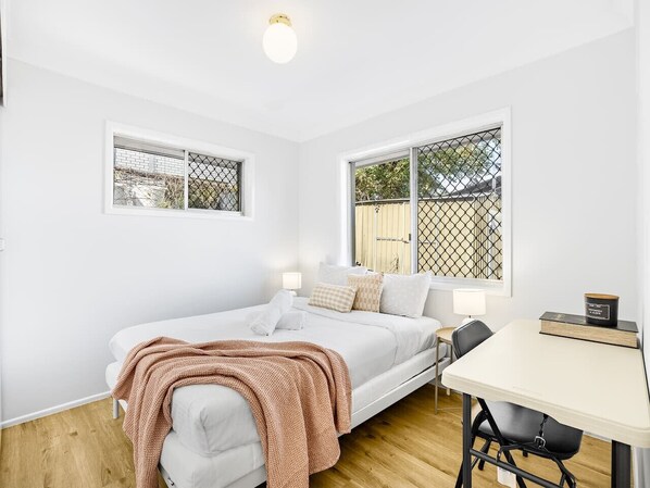 Una habitación pequeña y luminosa con una cama, una mesa y dos ventanas.