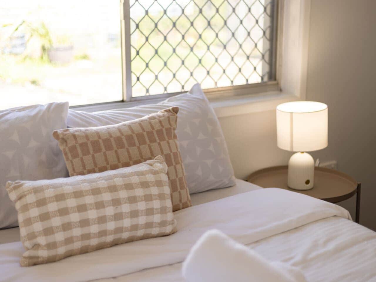 A bed with checkered pillows, a bedside lamp, and a window with a view of greenery.