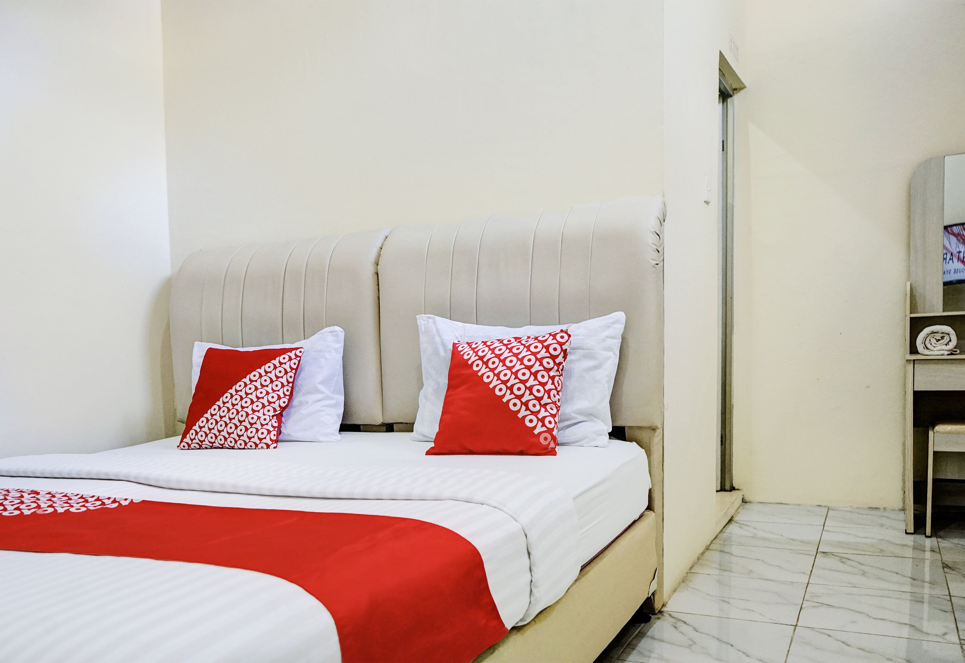Une chambre d’hôtel avec un lit, deux oreillers, une couverture rouge et blanche et une tête de lit blanche.