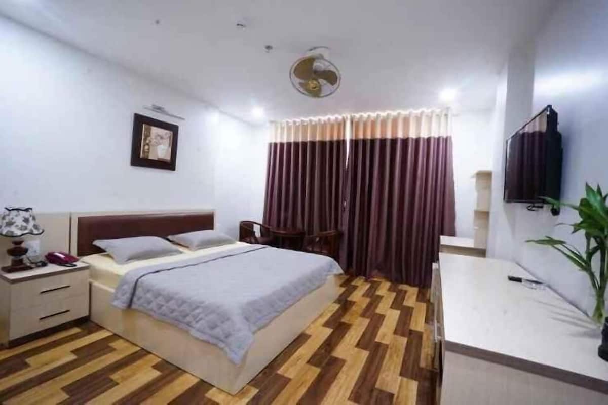 Une chambre à coucher avec un lit, une table de chevet, une lampe, des rideaux et un téléviseur.