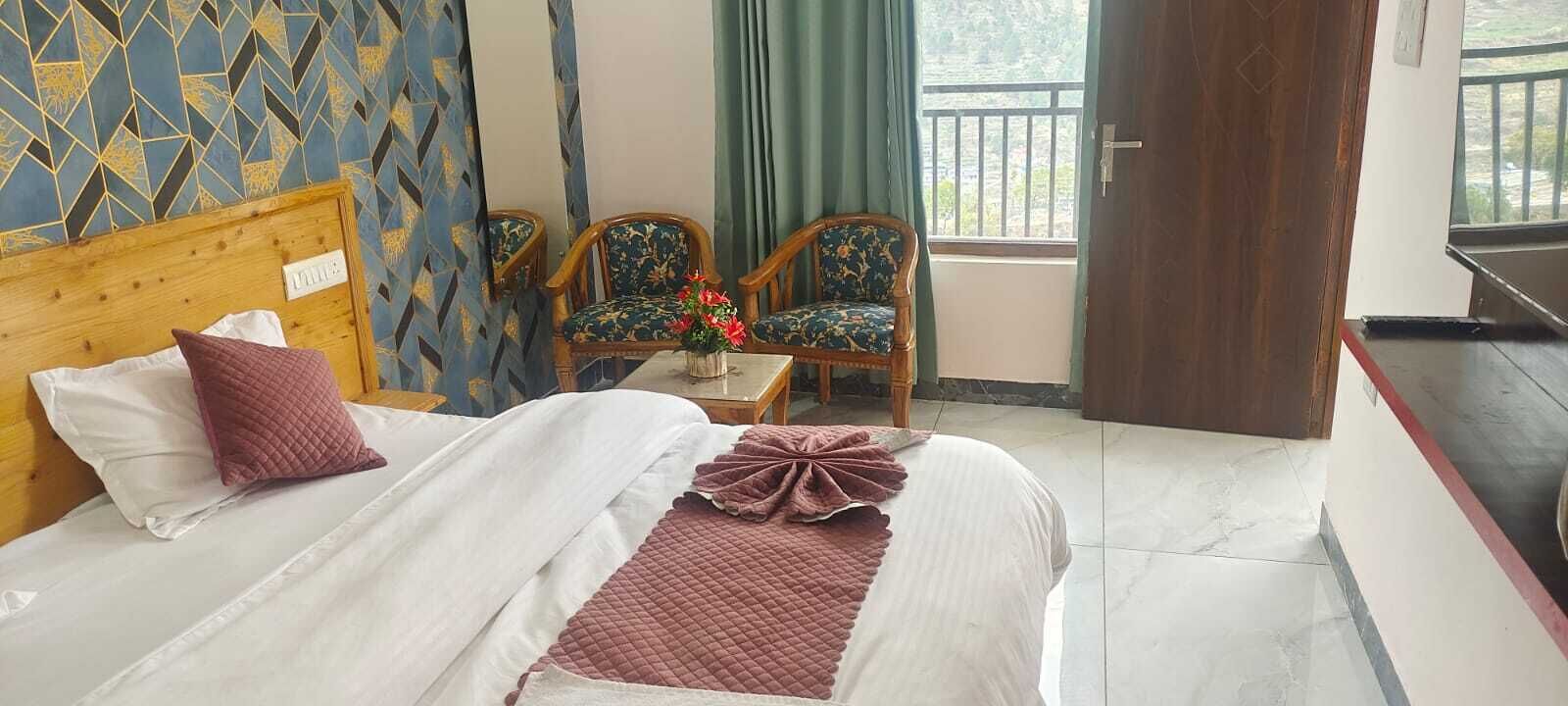 A hotel room with a bed, two chairs, a small table with a vase of flowers, and a view of the outdoors through a window.