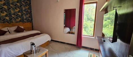 A hotel room with two beds, a TV, a small table, and a window with a view of greenery.