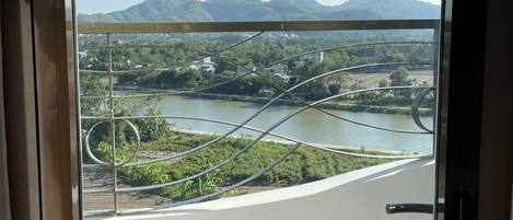A view from a balcony with a metal railing, overlooking a river, greenery, and distant mountains.
