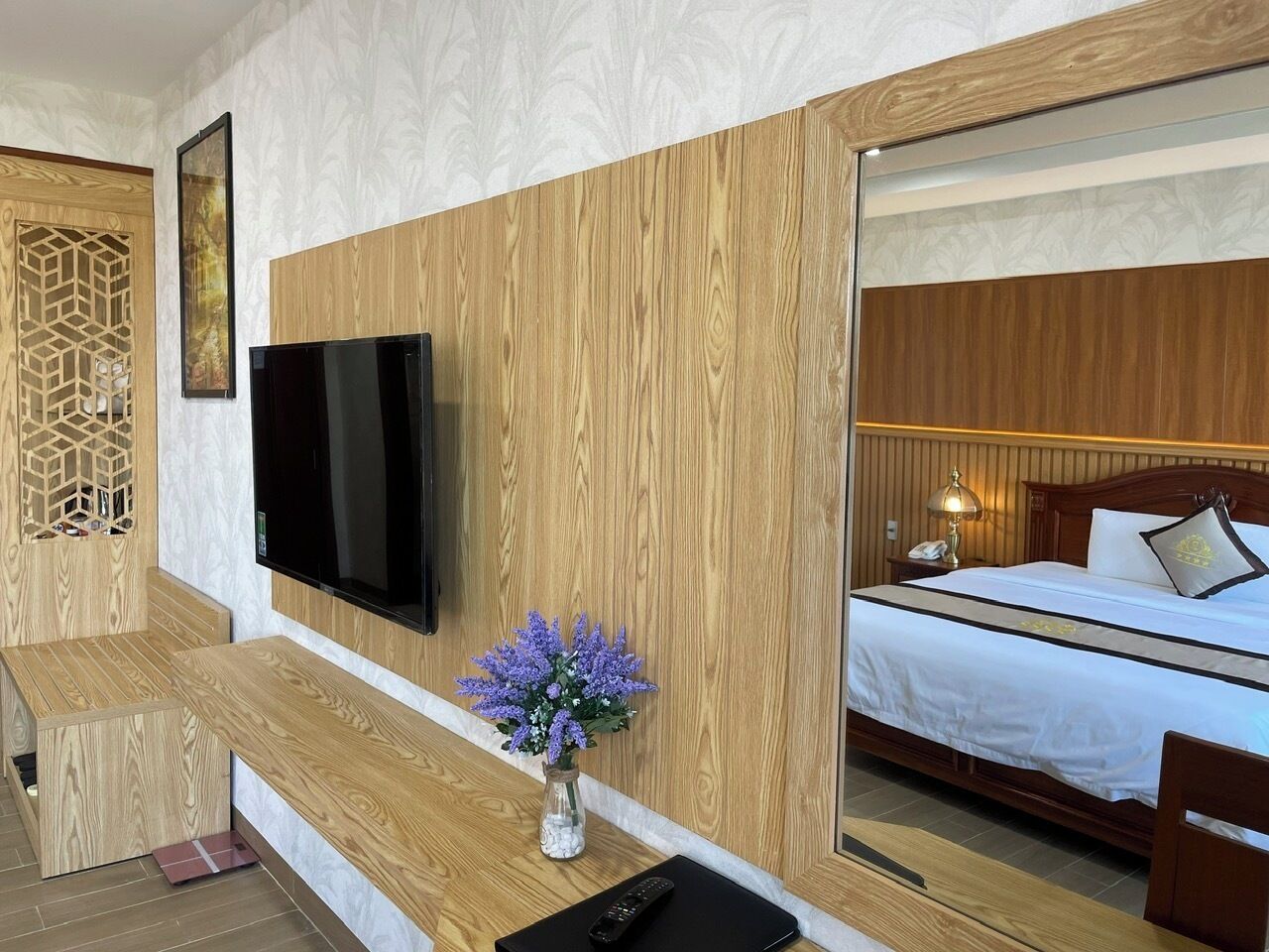 A hotel room with a wooden wall panel, a flat-screen TV, a bed with white bedding, a bedside table with a lamp, and a vase with purple flowers.