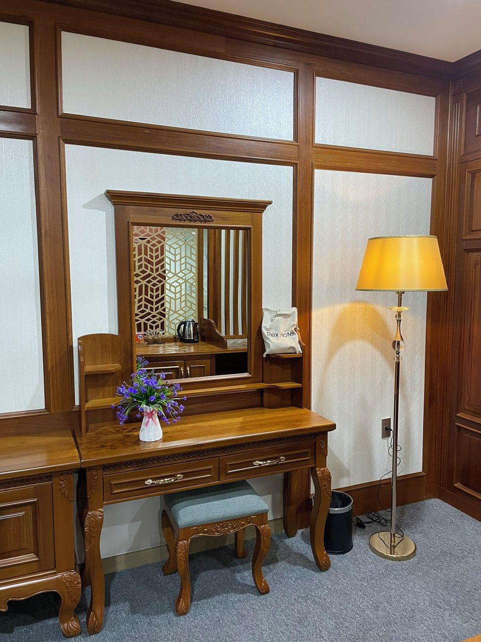 A wooden desk with a mirror, a lamp, and a vase with flowers.