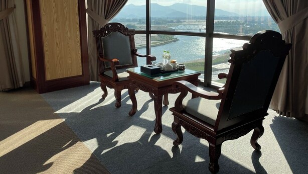 A room with a large window, two chairs, a small table with a bottle and a teapot, and a view of a bridge and mountains. - Rex Hotel Binh Dinh - by BAY LUXURY (Binh Dinh)