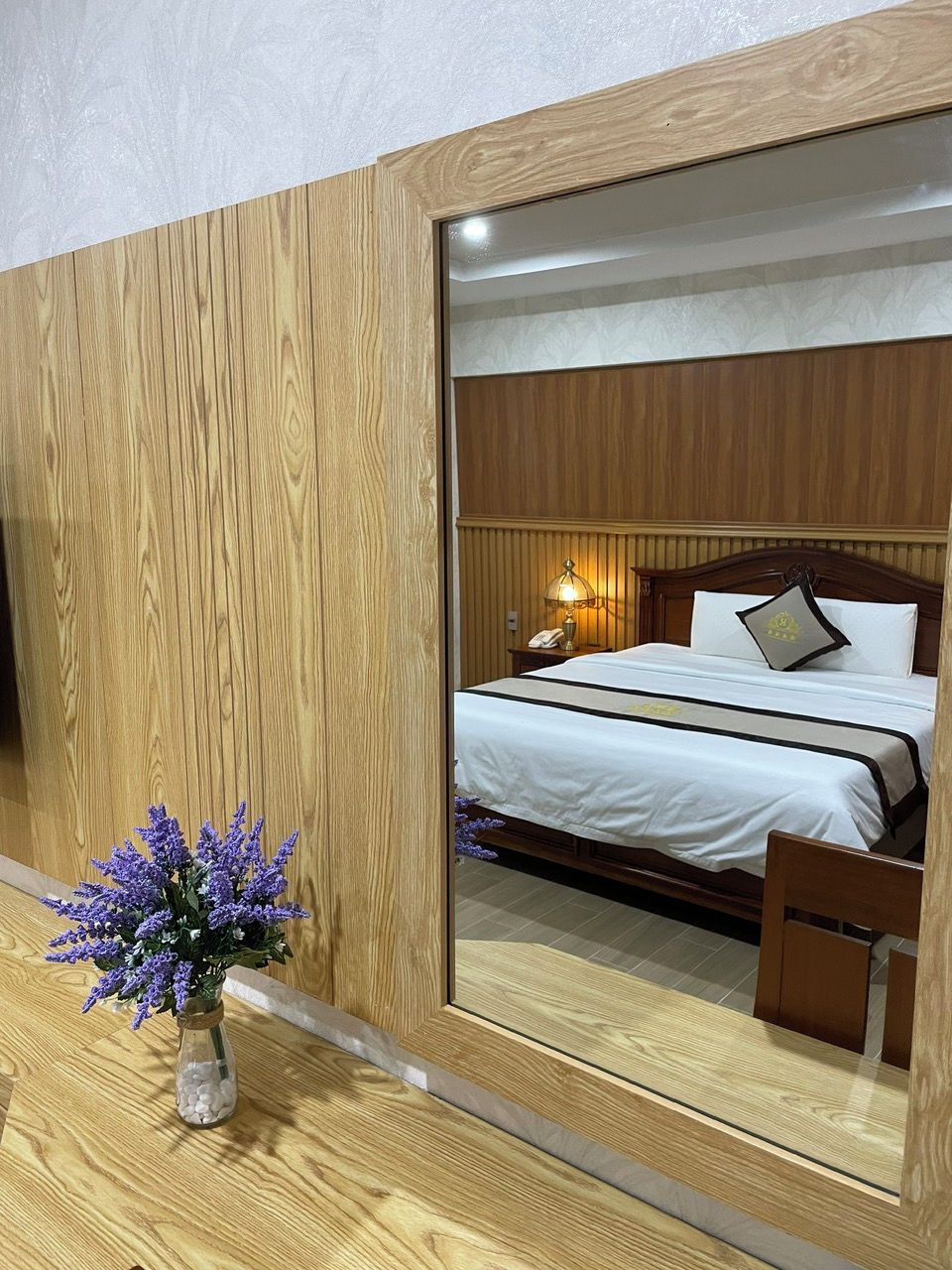 Ein Schlafzimmer mit einem Holzkopfteil, einem Bett mit weißer Bettwäsche, einem Nachttisch mit Lampe und einer Vase mit violetten Blumen auf einem Holztisch.