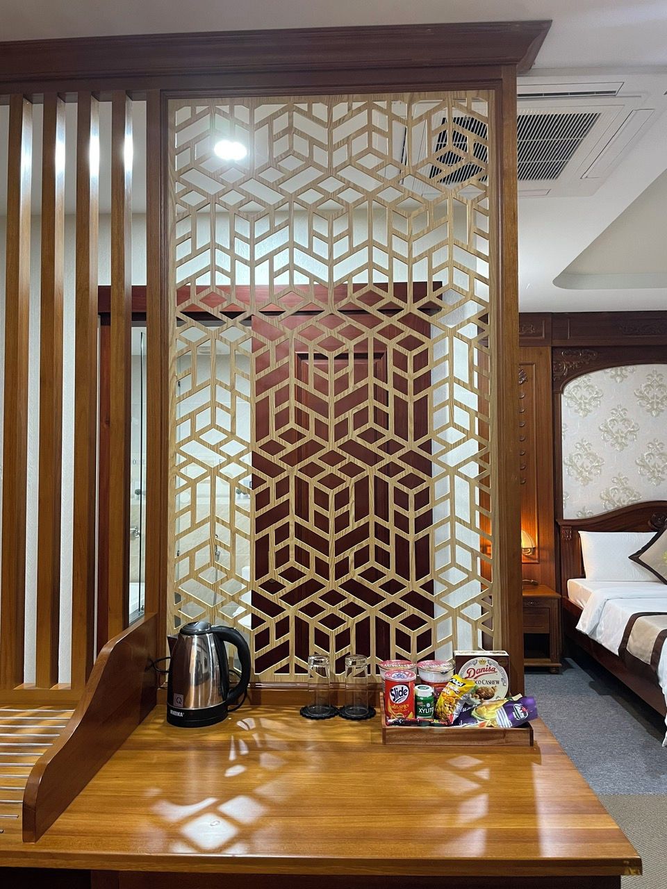 Camera d'albergo con un pannello divisorio in legno, un letto con biancheria bianca, un comodino con bollitore e snack, e una bocchetta di ventilazione a soffitto.