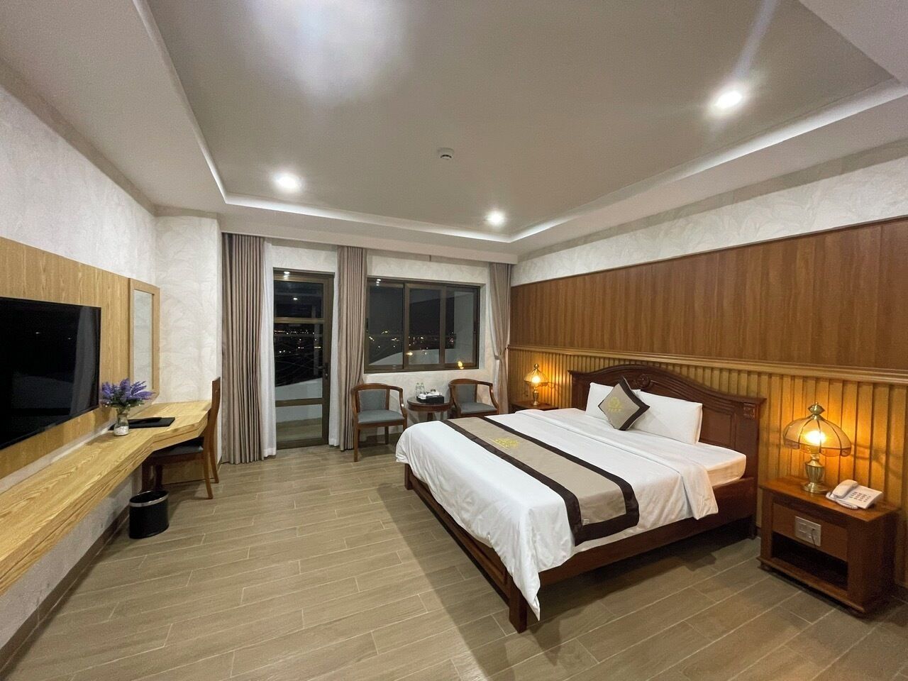 A hotel room with a large bed, a desk with a TV, a chair, a small table, and a window with curtains.