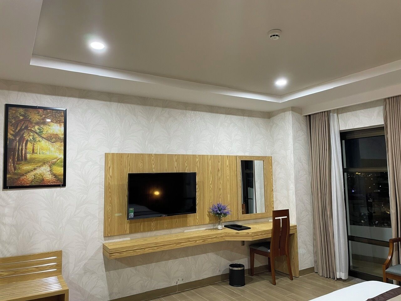 A hotel room with a wooden desk, a flat-screen TV, a mirror, a window with curtains, and a painting on the wall.