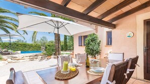 Un espace extérieur aménagé pour les repas, avec une table dressée pour deux personnes, un parasol blanc et une vue sur une piscine et de la verdure.