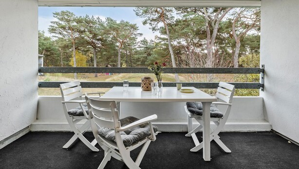 A covered patio with a table and four chairs, a vase with flowers, and a view of trees and grass.