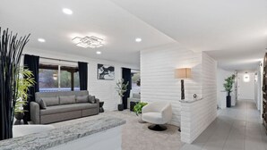 Un salon moderne avec un canapé gris, un fauteuil blanc et une table basse. On y trouve un mur en briques blanches, un lampadaire et une grande fenêtre avec des rideaux.