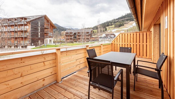 Una terraza de madera con una mesa y sillas, con vistas a una zona montañosa y edificios al fondo.