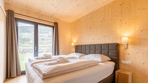 A bedroom with a large bed, wooden walls, a view of the outdoors, and two bedside lamps.
