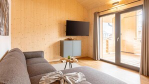A modern living room with a large flat-screen TV mounted on the wall, a grey sofa, a small wooden side table with a green mug, and a sliding glass door leading to an outdoor wooden deck.