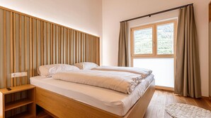 A bedroom with a wooden bed, a bedside table, a window with curtains, and a view of greenery outside. - Beautiful apartment with balcony (Kreischberg Murau)