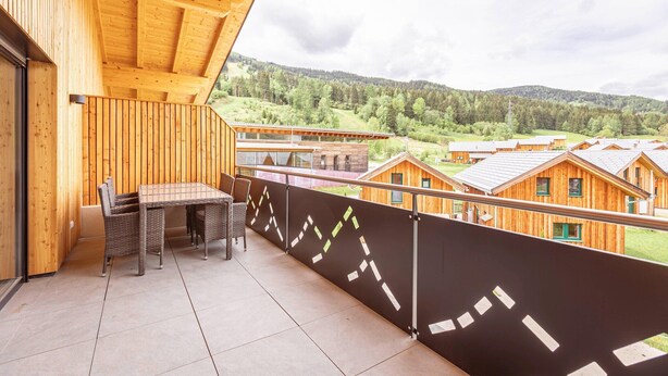 A wooden balcony with a table and chairs, overlooking a forested valley with multiple wooden cabins. - Beautiful apartment with balcony (Kreischberg Murau)