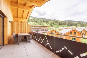 Ein Holzbalkon mit Tisch und Stühlen, mit Blick auf ein bewaldetes Tal und mehrere Holzhütten.