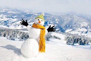 Un muñeco de nieve con sombrero verde y bufanda amarilla en un paisaje nevado con montañas al fondo.