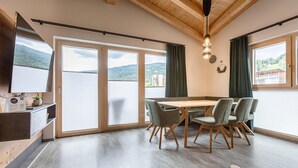 Zona pranzo moderna con tavolo e sedie in legno, ampie finestre con tende e vista su un lago e montagne.